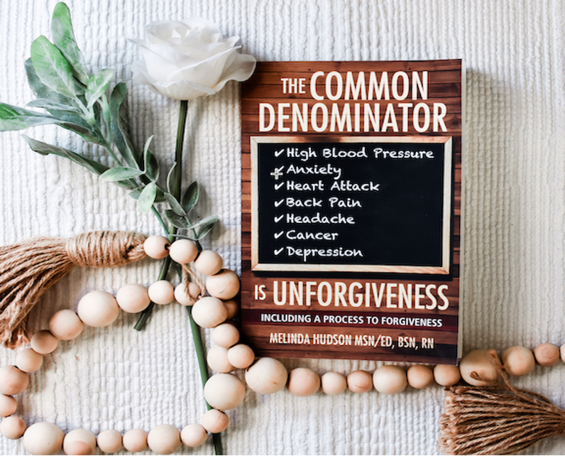 The Common Denominator Is Unforgiveness by Melinda Hudson displayed with a white rose and wooden beads